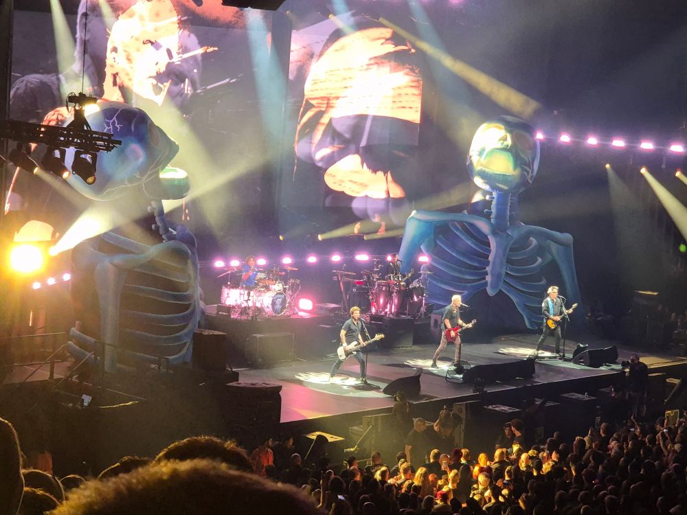 A stage with the members of the band Offspring playing, with giant inflatable skeletons at stage rear and a screen behind showing closeups of Dexter, the lead singer