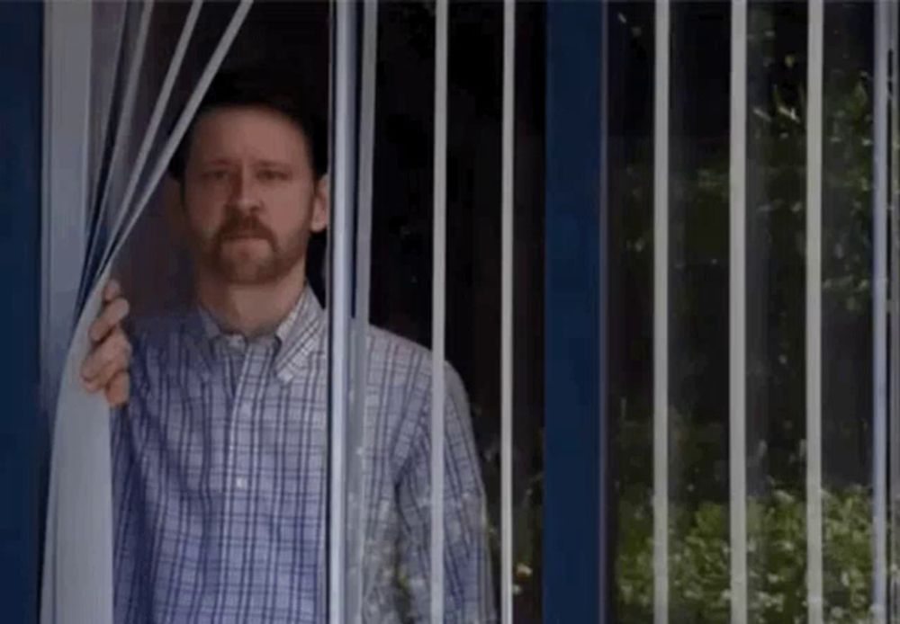 a man in a plaid shirt is standing in front of a window behind bars .