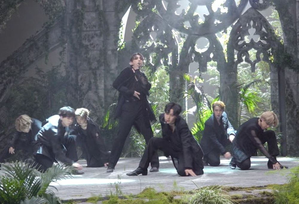 Screencap of BTS's performance of Black Swan; they're on a stage backed by what looks like the ornate window of a ruined church with pale light streaming through it, with greenery behind and around them. They're dressed in witchy black. JK is in the centre singing, and the others are half-kneeling picturesquely around him