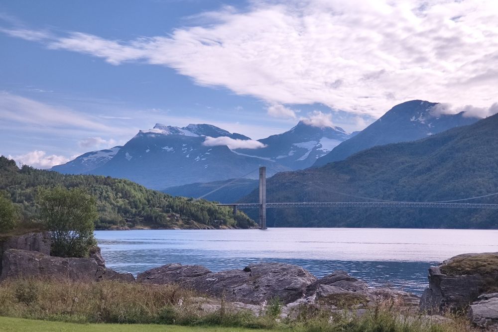 Norwegian fjord in the foreground, Skjombrua bridge midground and mountains with wispy clouds and patches of snow in the distance.