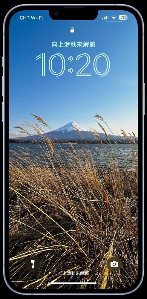 This image shows an iPhone lock screen displaying a scenic wallpaper. The wallpaper features golden grass in the foreground with a clear view of Mount Fuji in the background under a blue sky. The iPhone frame is visible, and the lock screen elements include the time, "swipe up to unlock", and icons for flashlight and camera at the bottom.