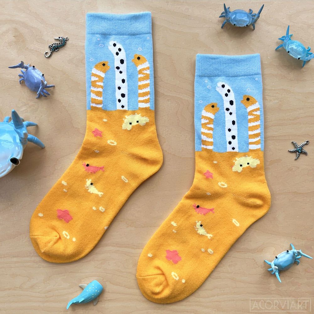 photo of a pair of socks laid flat. the socks have garden eels peeking out of sand above the ankle area, with shrimp, crabs, and starfish scattered around lower on the foot. small crab pen holders are used as props