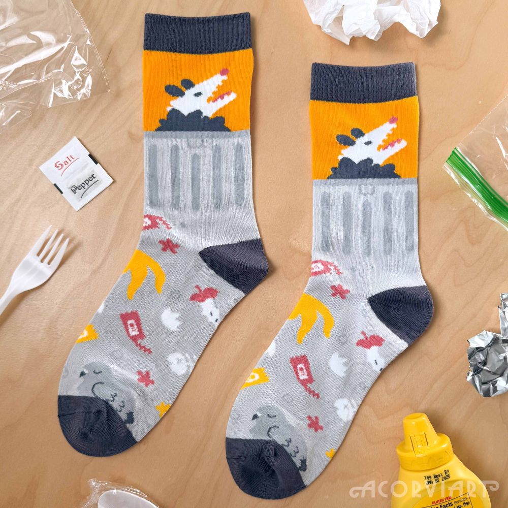 Socks that have a city trash theme, with a pattern of banana peels, eggshells, and condiment packets on the main body. The ankle of the sock is a trash can, with an opossum poking its head out the top of the can. A pigeon sleeps at the foot of the sock.