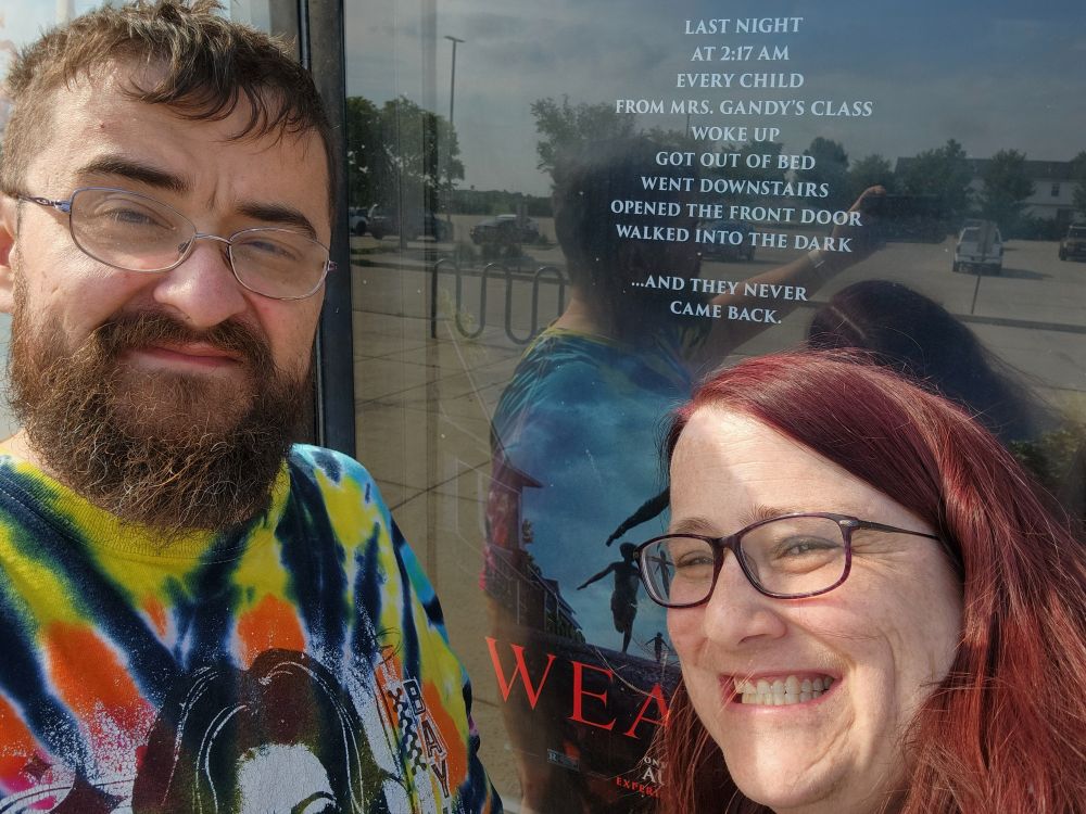 Me and my spouse in front of the Weapons (2025) poster.