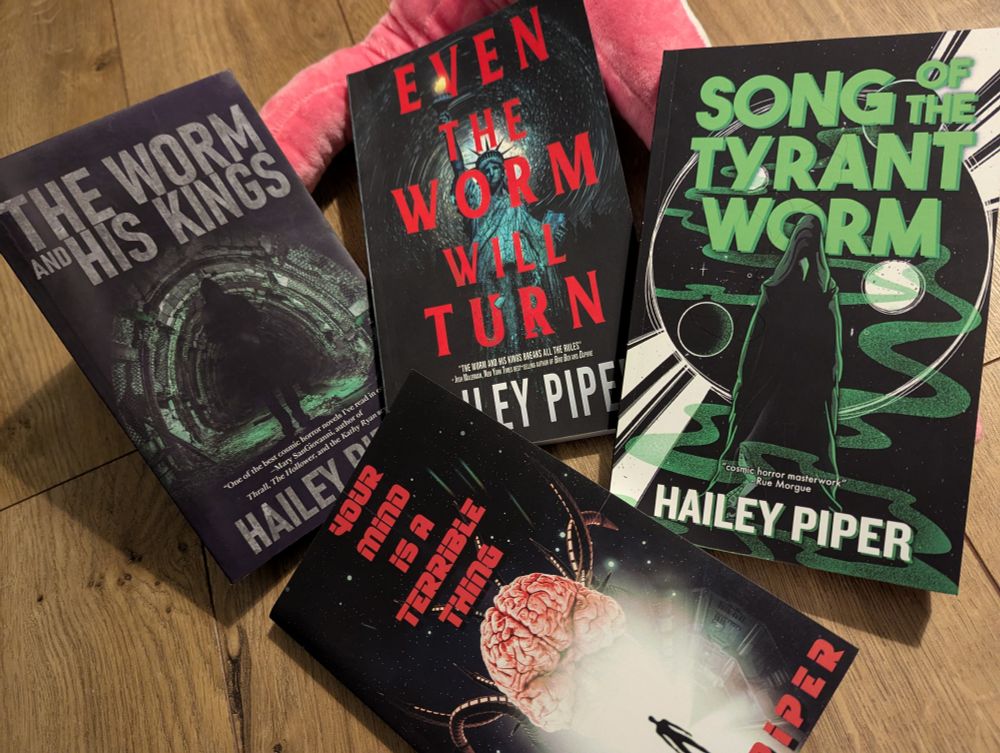 a pile of books by Hailey Piper, showing The Worm and His Kings, its sequel Even the Worm Will Turn, the trilogy's conclusion, Song of the Tyrant Worm, and a standalone book Your Mind is a Terrible Thing 