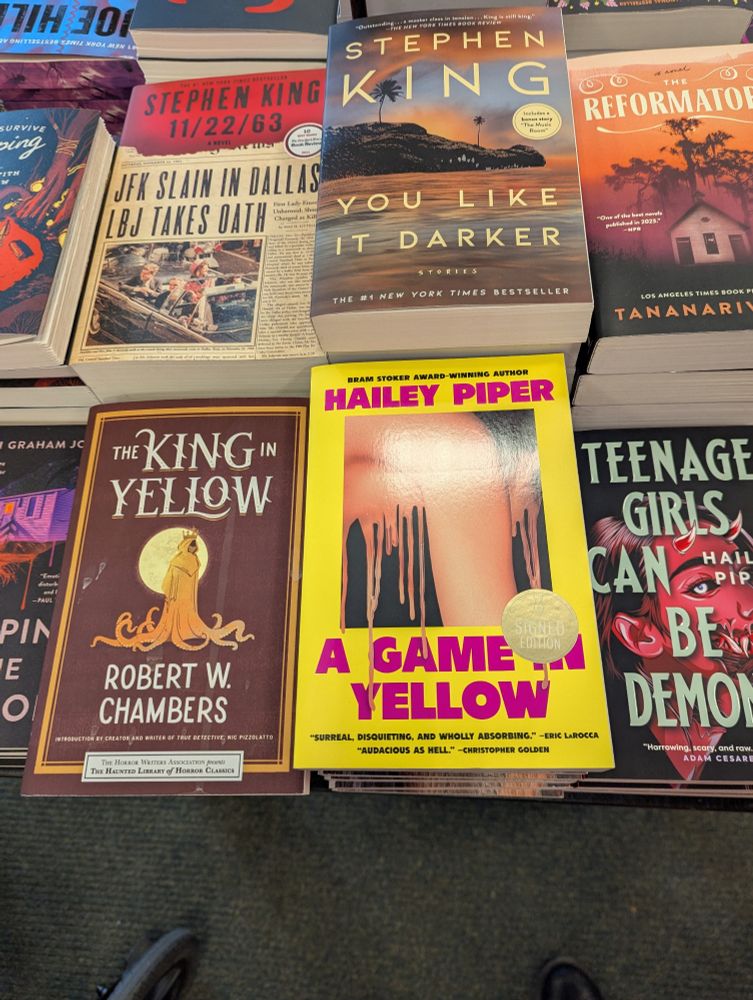 A Game in Yellow by Hailey Piper, art by Caroline Johnson, next to The King in Yellow by Robert W Chambers among different horror books by Tananarive Due, Stephen King, and more