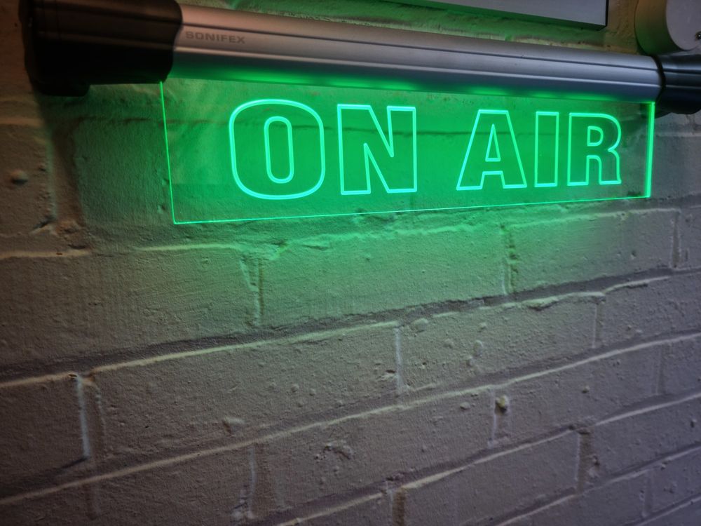 A lit neon green sign against a white brick background. The sign read 'On air' to indicate filming is in progress.  