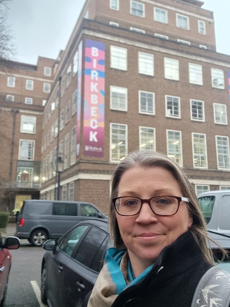 A smiling middle-aged aged woman standing in front of Birkbeck, University of London, buildings, ready for a day of presentations about immersive learning. 
