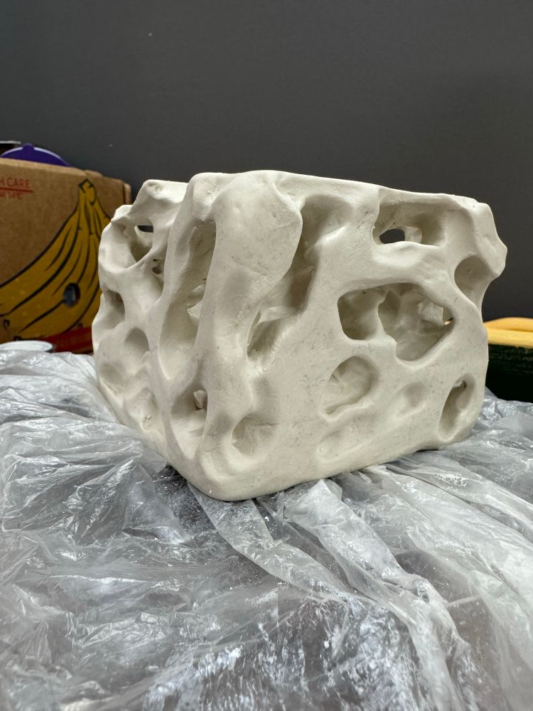 Side, corner view of a greenware porcelain cube with carved holes to look like eroded rock. It’s sitting on plastic on a cluttered work table.