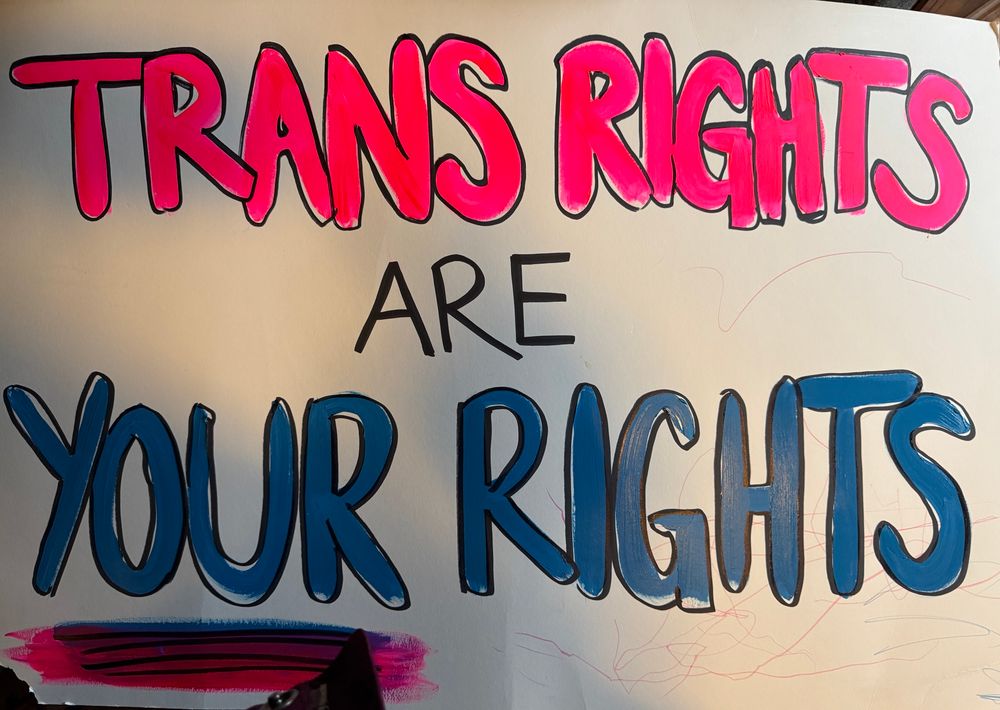 Protest sign painted in neon pink and blue bubble letters that says “TRANS RIGHTS ARE YOUR RIGHTS.” “YOUR” is underlined.