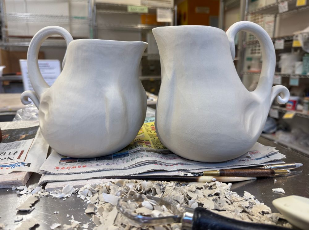 Two greenware porcelain pitchers sitting on newspaper on a worktable in a pottery studio. The pitchers almost look like two birds facing each other with the handles like tail feathers and the mouths like beaks.