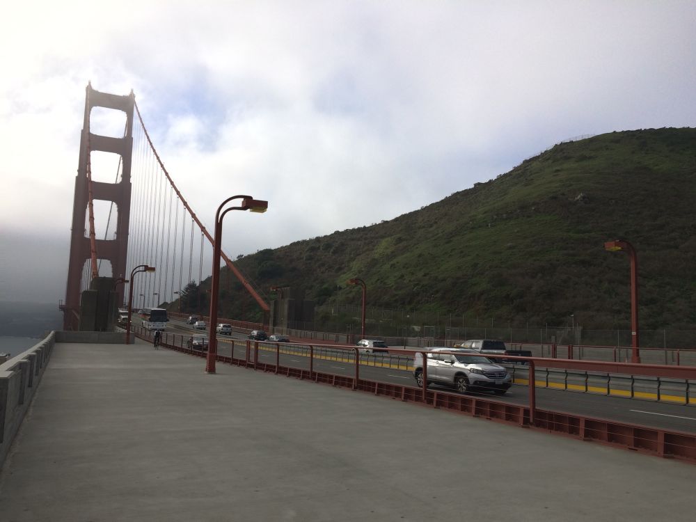 An oblique photo of the Golden Gate Bridge from the Marin side. The roadway curves along to the right of the frame in front of a green hill, while the bridge rises along the left side and nearly disappears into fog.