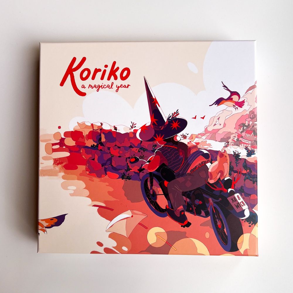 The cover art of Koriko: a magical year. Featuring a person on a motorcycle with a cat riding on the back. The person is wearing jeans covered in patches and a tall witch's hat. A cat sits on the back of the bike and the view over the stone wall shows a town on a seaside cliff.