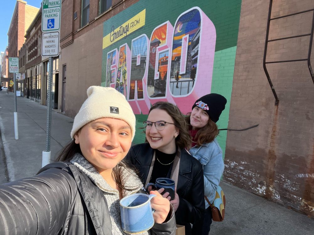 Three people in front of a “greetings from Fargo mural” holding coffee mugs