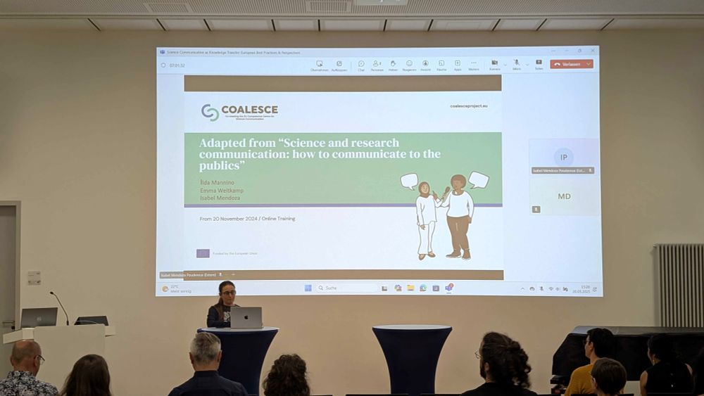A person standing behind of a laptop on a lecturn, in front of a presentation screen. Some heads of audience members are visible in the foreground.