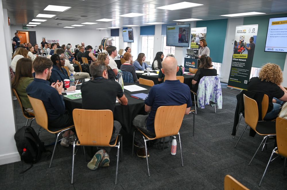 A large group of people sat around tables listening to a presenter talking from the front of the room