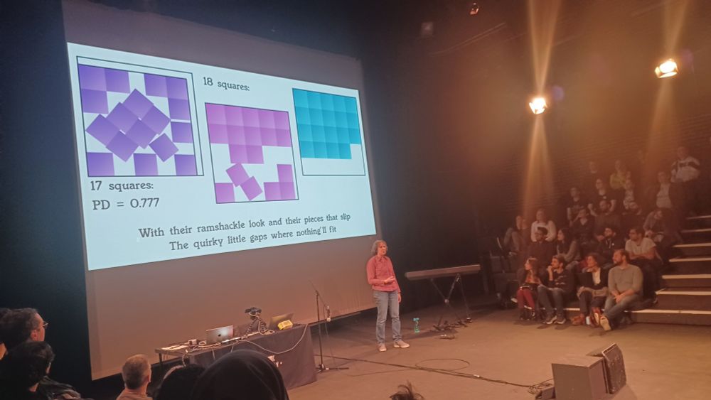 Photo of me on stage with square packing for 18 squares and 19 squares inside a bigger square on slide behind. The slide includes song lyrics 'With their ramshackle look and their pieces that slip, The quirky little gaps where nothing'll fit'. A few audience members are also visible.