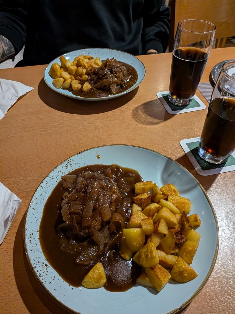 Zwei Teller stehen auf einem Tisch neben zwei Gläsern Cola. Auf den Tellern befinden sich jeweils ein veganer Hackbraten aus Nuss mit dunkelbrauner Soße mit Zwiebeln, daneben Bratkartoffeln 