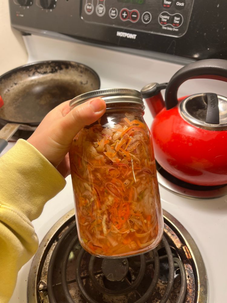 Jar of curtido made with red onions, cabbage, and carrots. Held in hand in front of stove top.