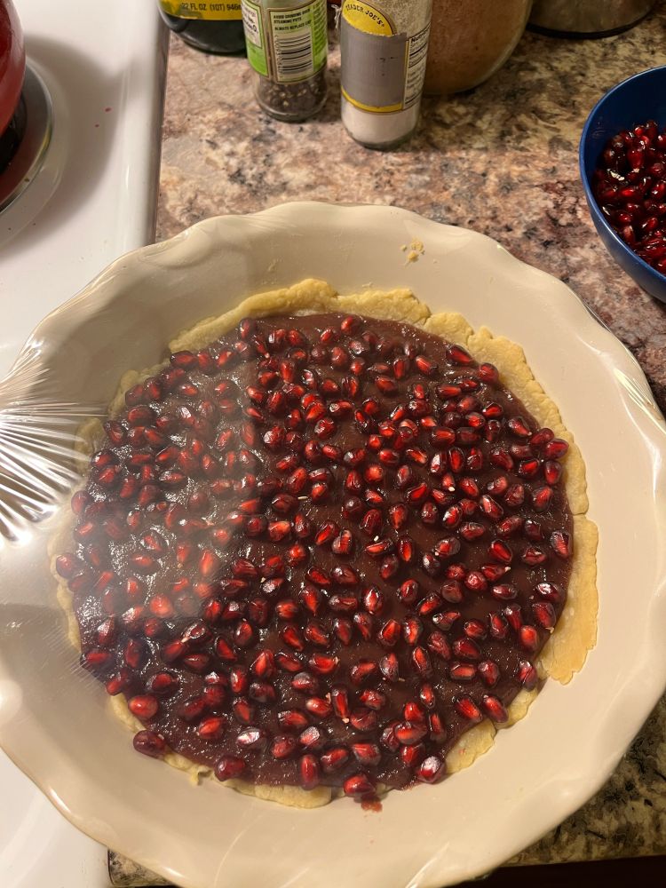 Pomegranate seeds scattered on a cranberry pomegranate tart inside a pie dish. There is plastic wrap cover the dish creating a glare on the left side of the dish.