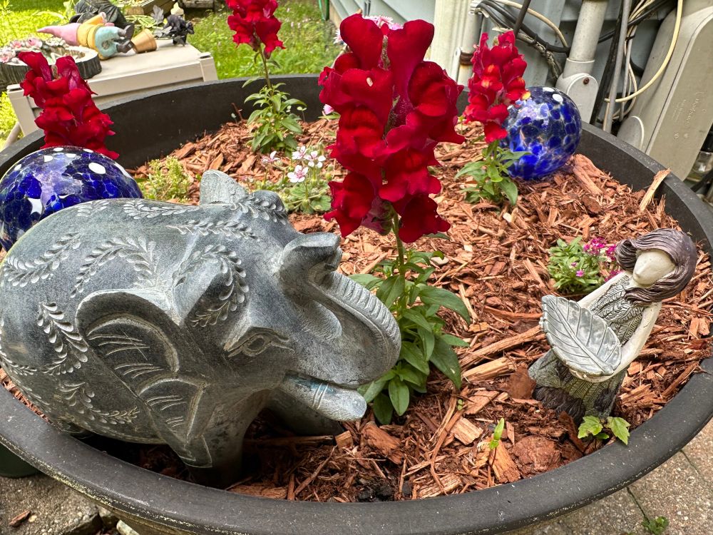 Photo of a flowerpot containing flowers, glass balls with swirls of blue and white, a stone elephant, and a little stone woman holding a leaf out towards the elephant.