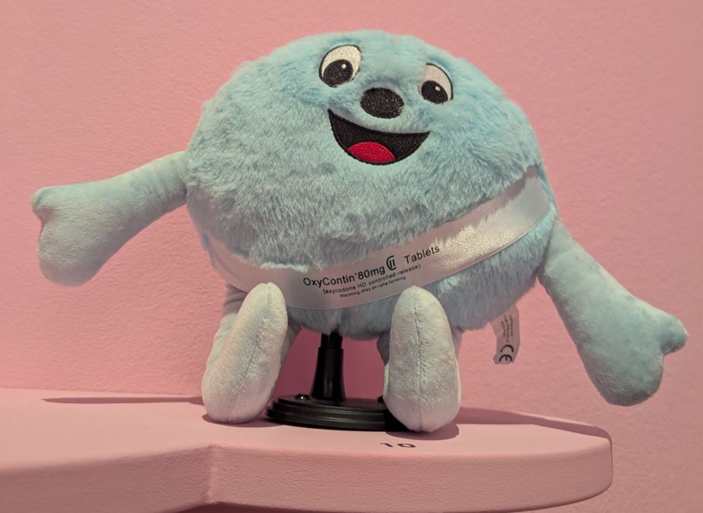 A blue stuffed animal but it's actually a smiling Oxycontin pill, on a pink background 