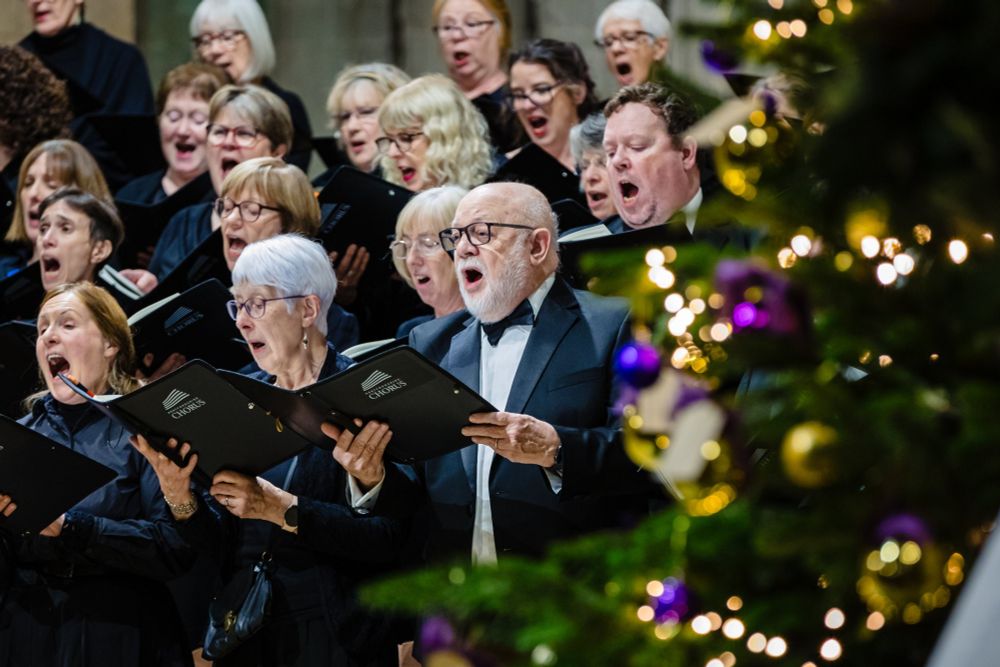 A choir dressed in black gowns and dinner jackets hold black folders and sing. In the foreground is a Christmas tree decorated with gold and purple ornaments.