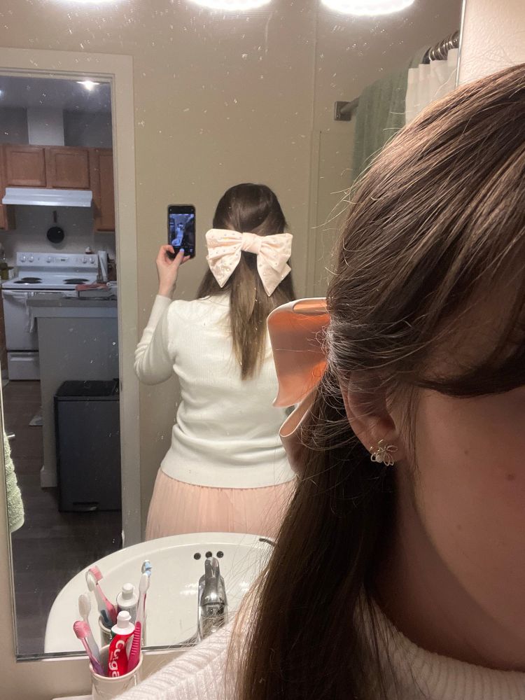 A selfie that leverages a mirror so you can see the big, pastel pink sequined bow pinned to the back of the hair. The outfit is a white turtleneck with a matching pink skirt. 