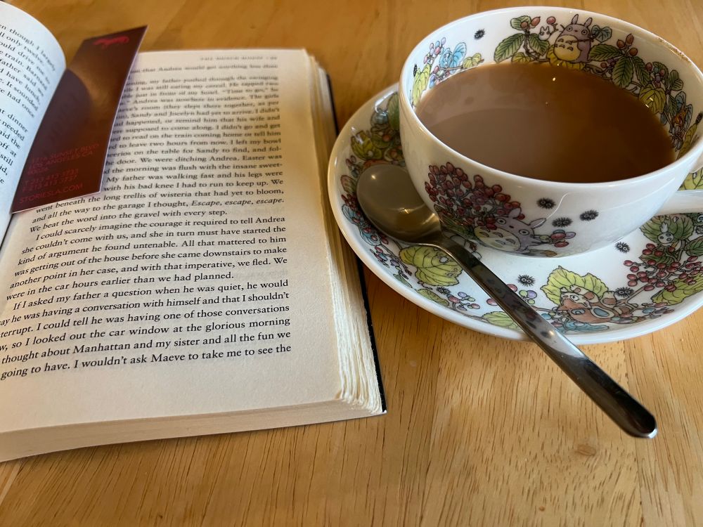 To the right, a Totoro tea cup filled with royal milk tea, and a stirring spoon. To the left, an open book with a bookmark between the pages. 