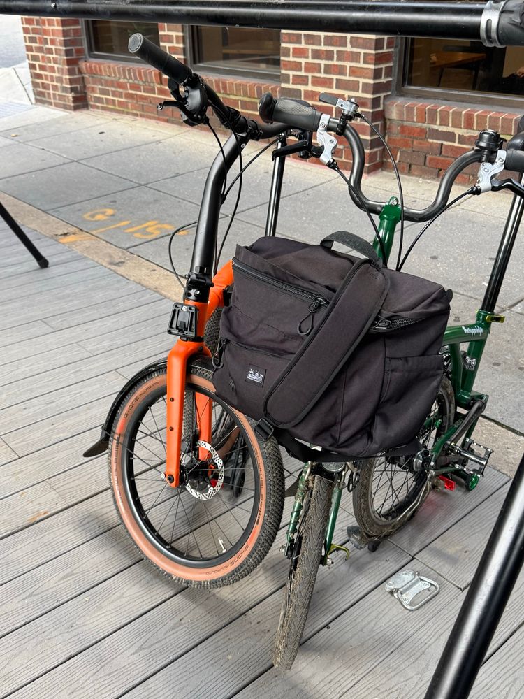 Orange Brompton folded next to green Brompton that has a bag on the front of it. In front of a bike stand