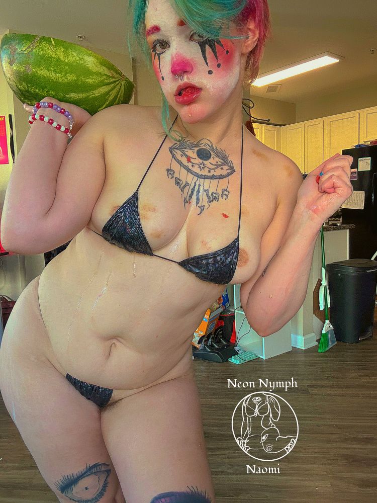 Naomi is holding half a watermelon in one hand and eating a piece they tore out of it with the other you can see they are drenched in watermelon juices in their black holographic micro bikini they also have a clown makeup look with the lower half gone from eating the watermelon their blush is pink that fades into orange and then yellow on a white base with asymmetrical graphic clown eyeliner