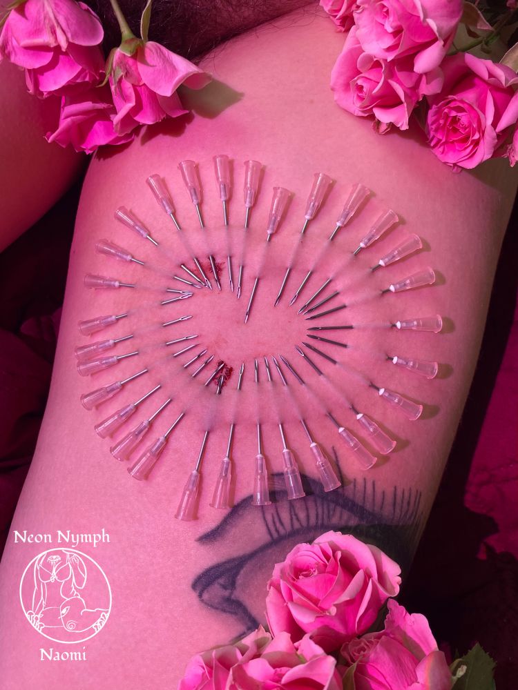 In the photo you see Naomi’s thigh in pink lighting with pink roses surrounding a needle play pattern in the shape of a heart you can they used 30 needles to make the heart shape in Naomi’s skin. 