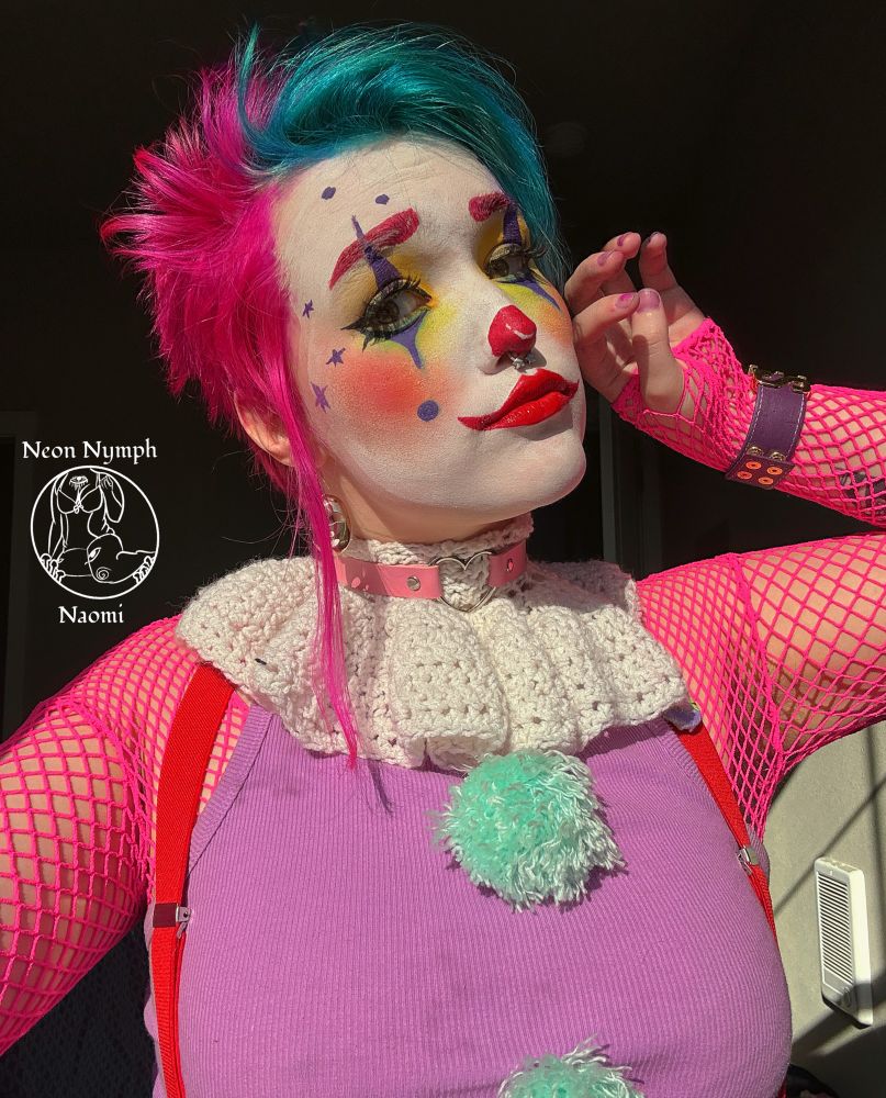 Naomi has hot pink hair with blue bangs they have clown makeup with a white base a red spikey lip in the shape of a smile.  They have red blush that blends into green shadow that’s defused under the eye they also have a cat eye eyeliner. They also have purple spikes under and on top of their eyes that has a purple dot at the bottom of them.  They are wearing a neon pink fishnet top with a light purple halter top that has bright blue pompoms on it.  They are looking at the camera that is at a lower angle and their hand is pressed to their cheek.  