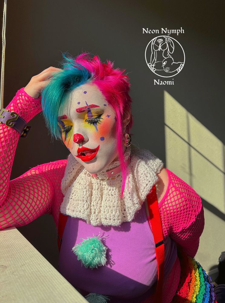 Naomi has hot pink hair with blue bangs they have clown makeup with a white base a red spikey lip in the shape of a smile.  They have red blush that blends into green shadow that’s defused under the eye they also have a cat eye eyeliner. They also have purple spikes under and on top of their eyes that has a purple dot at the bottom of them.  They are wearing a neon pink fishnet top with a light purple halter top that has bright blue pompoms on it.  They are looking at down and their hand is messing with their hair. 