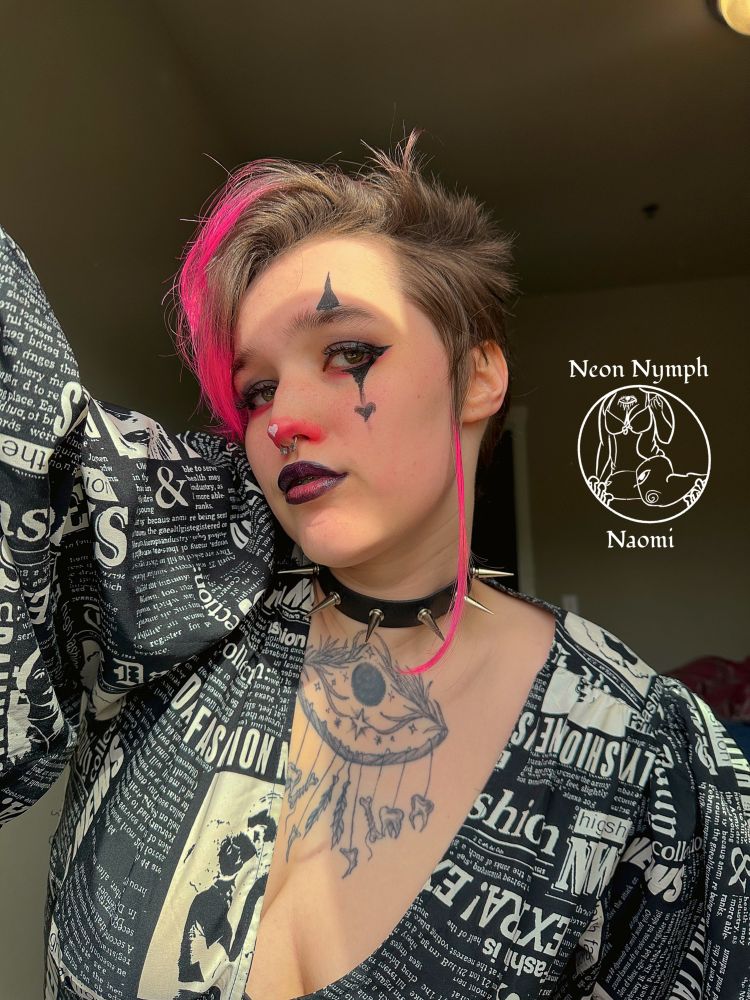 Naomi has on a cropped shirt with newspaper clipping design all around it it’s a shirt with big bubble sleeves and a deep v neckline. They are also wearing a black leather spiked choker. They also have neon pink dyed ends to their brown hair.  Their makeup consists of: a red nose with a white heart in the middle, a black Smokey eye with red on the bottom that’s smudged out, a black glossy lip with a red center on the bottom lip, they also have clown graphic liner with a heart at the bottom of the bottom spike. 