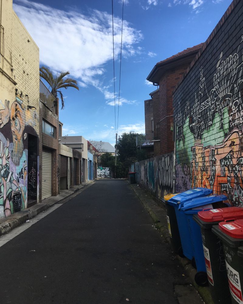 Looking down a lane in suburban Enmore, Sydney. The sky above is blue with a few white wispy clouds. The back of buildings on both sides are covered in colourful graffiti. On the right there is a row of 5 Otto bins with yellow, blue and red lids