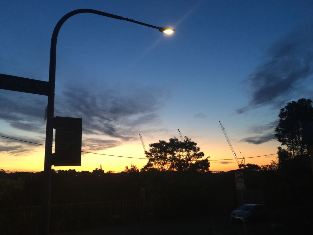 Photo of sunset, with the sky changing from dark blue to light blue to orange where it meets the horizon. On the horizon is a silhouette of a large tree and two cranes further back. In the foreground on the left a streetlight post rises and curves so the streetlight hangs in the centre top of the view