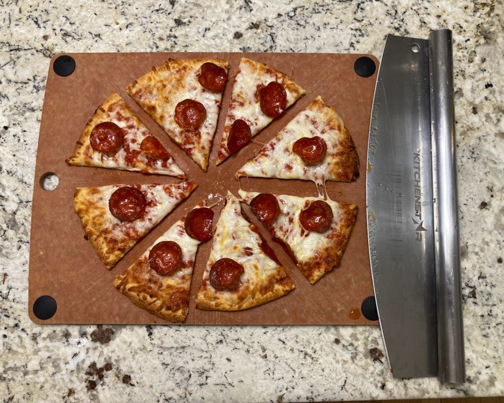 A small, pepperoni, cauliflower-crust pizza sits on a cutting board, having been cut cleanly into 8 sectors. Beside the pizza sits what is essentially a large, rocking knife with a handle running along the entire length of the blade. 