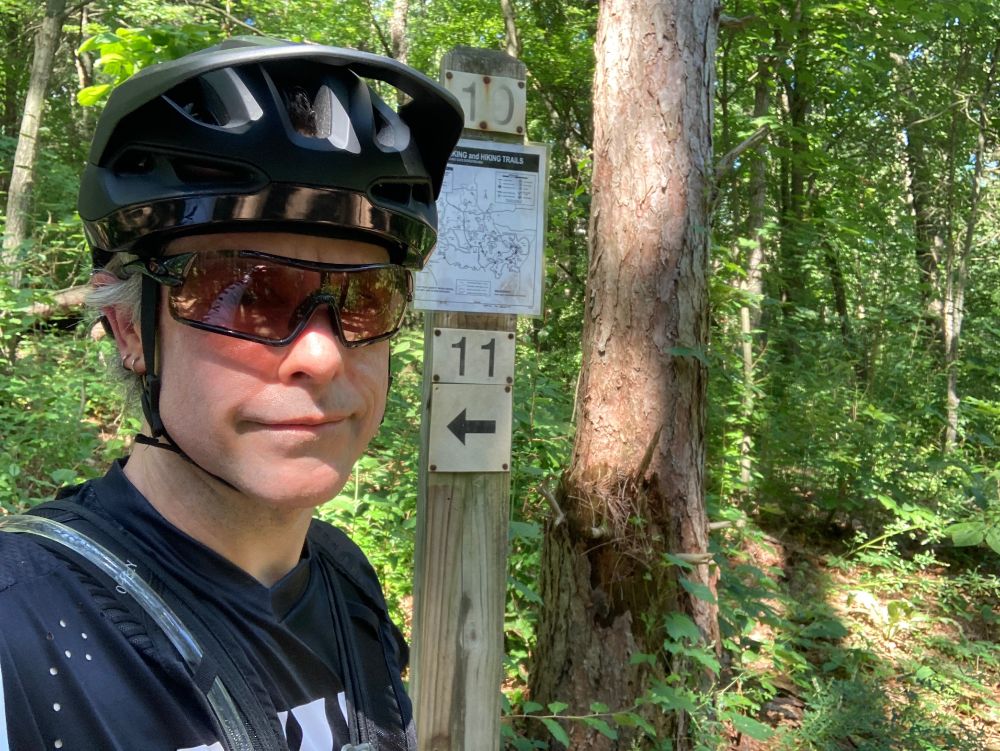 A human with light skin and gray hair stands in black/white cycling gear in front of a trail marker.