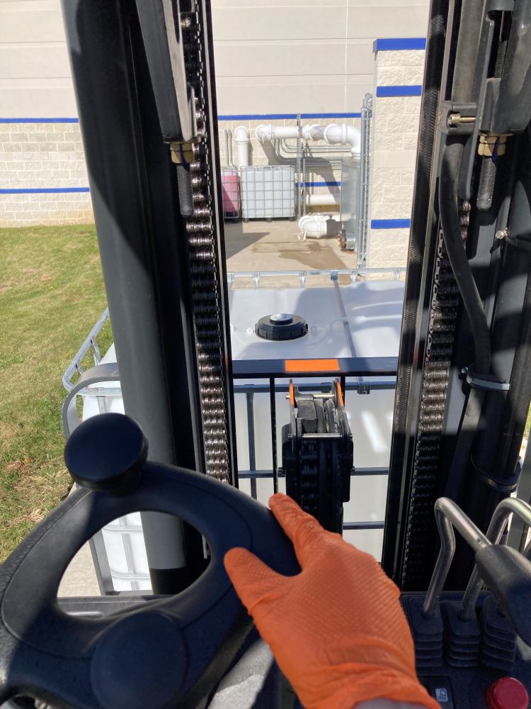 A photo from the operator's seat on a fork truck shows an orange-gloved hand on the oblong steering wheel and an empty 275-gallon tote on the truck's fork. Another empty tote site neatly beside a full one in the distance.