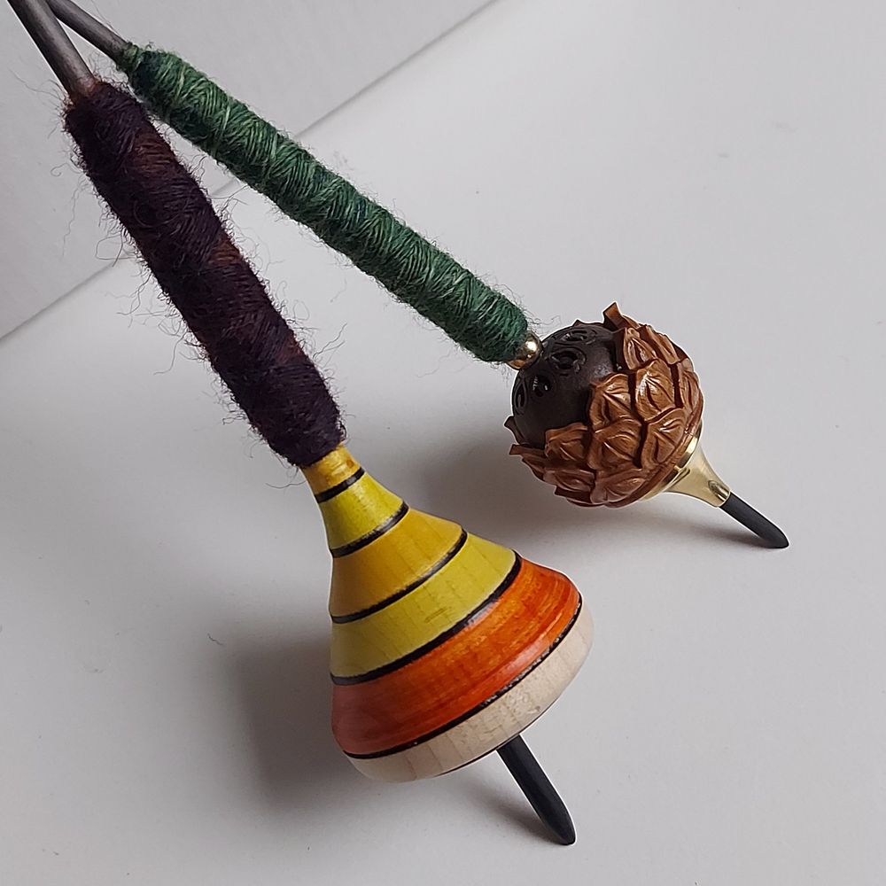Closeup of two spindles, both with a black  carbon fiber shaft, are leaning against a white box on a white table.
For the front spindle a yellow and orange striped spinning top was used, for the back one a carved bead from a Mala.
Both are filled with some fiber samples, in front in red and violet tones, in the back with greenish tones.

#spinning
#handspinning
#handspun
#supportedspinning
#spinnersofsky
#spinsky
#fiberarts
#fiber
#wool
