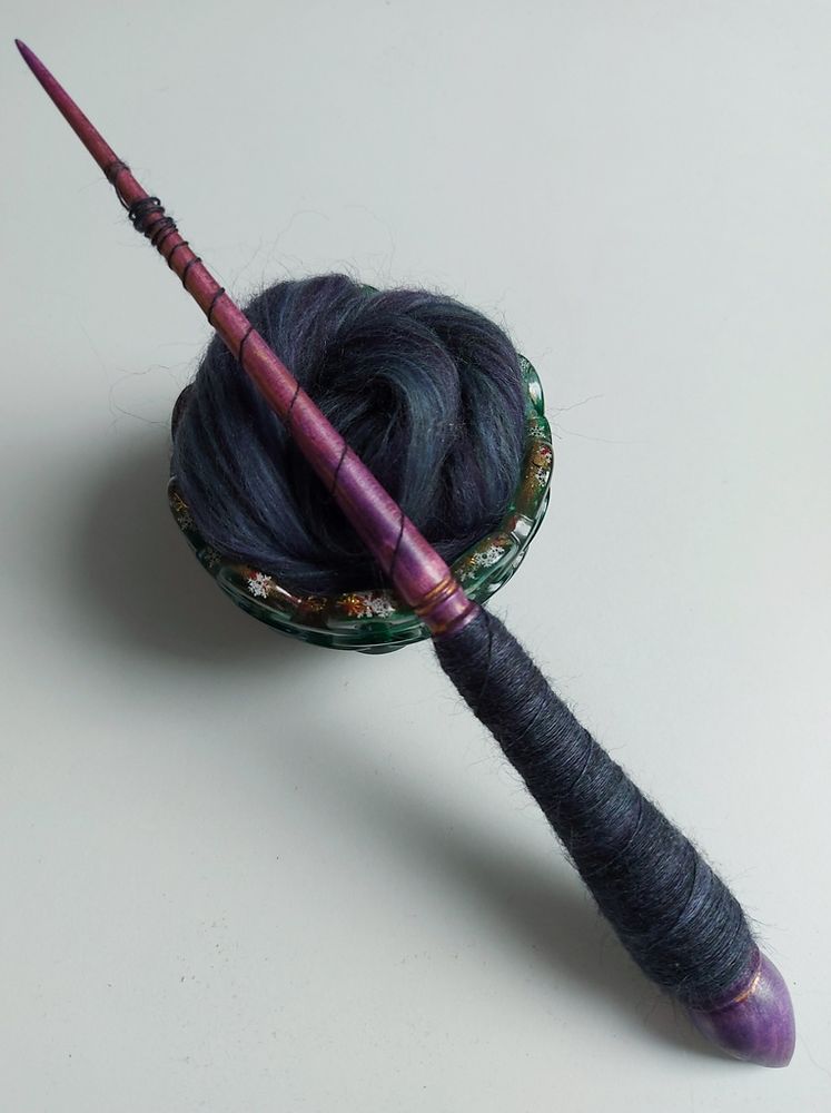 a violet colored, half filled, wooden support spindle, laying on a green spinning bowl filled with very dark fibers with green, violet and blue shades
