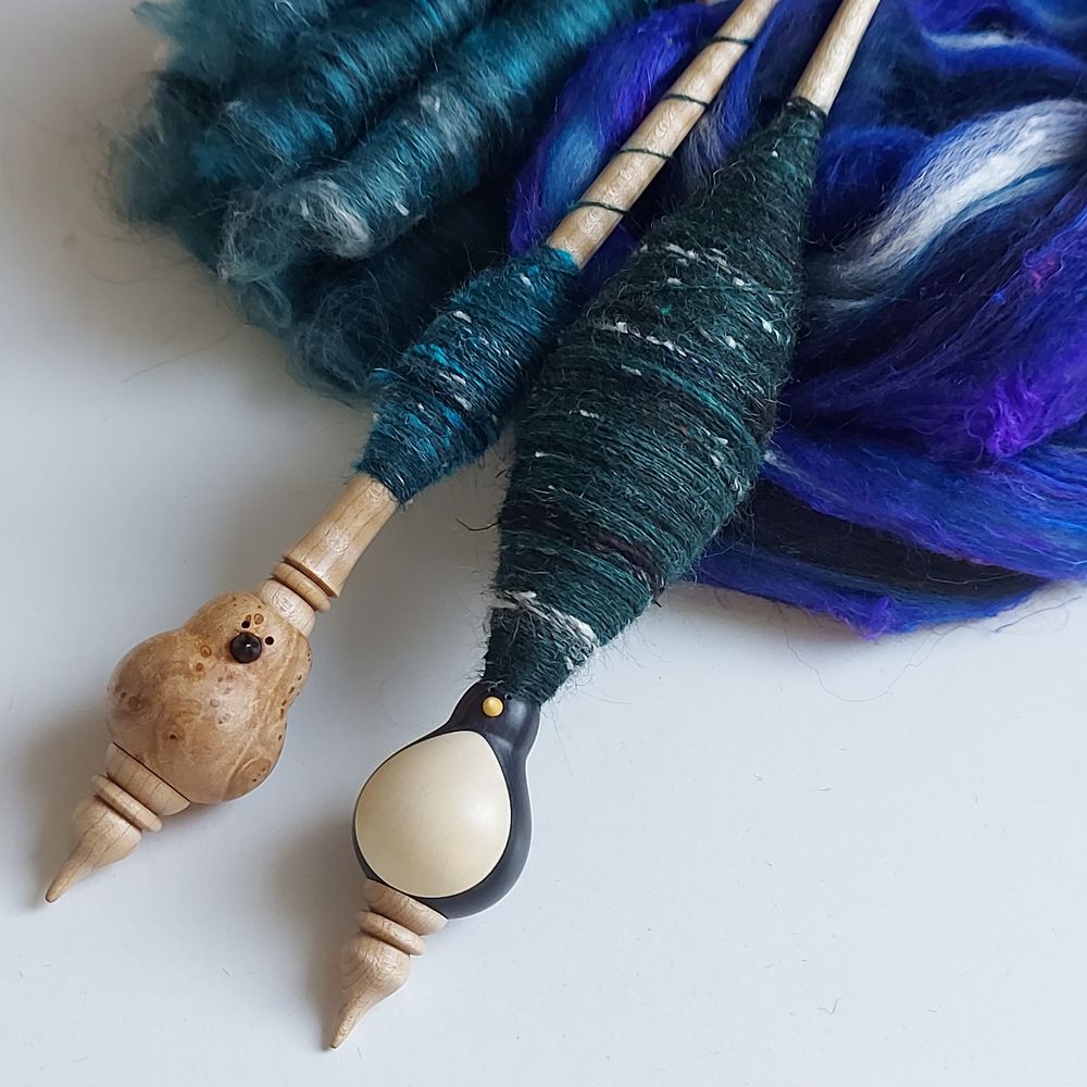 Two wooden, bird shaped supported spindles are laying on fibers with different shades of blue and green. Fibers in the left upper corner are formed into rolags to get a more mixed colorway.