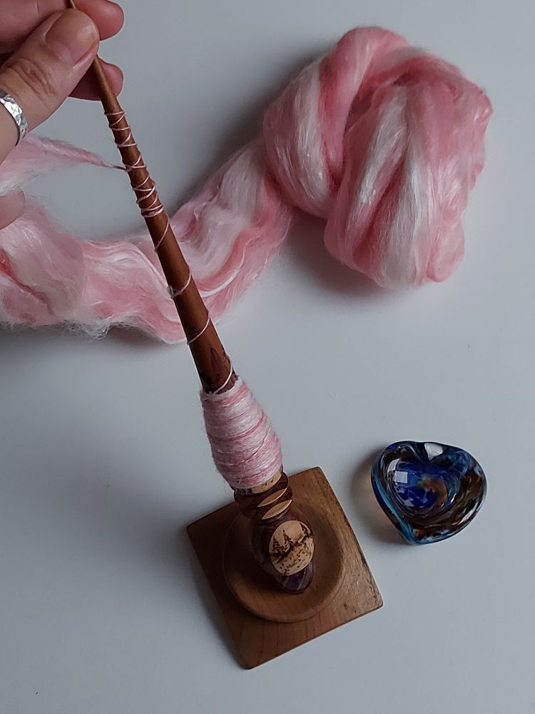 a supported spindle in a wooden bowl surrounded with pastel pink and white Polwarth fiber, next to it a heart shaped glass bowl