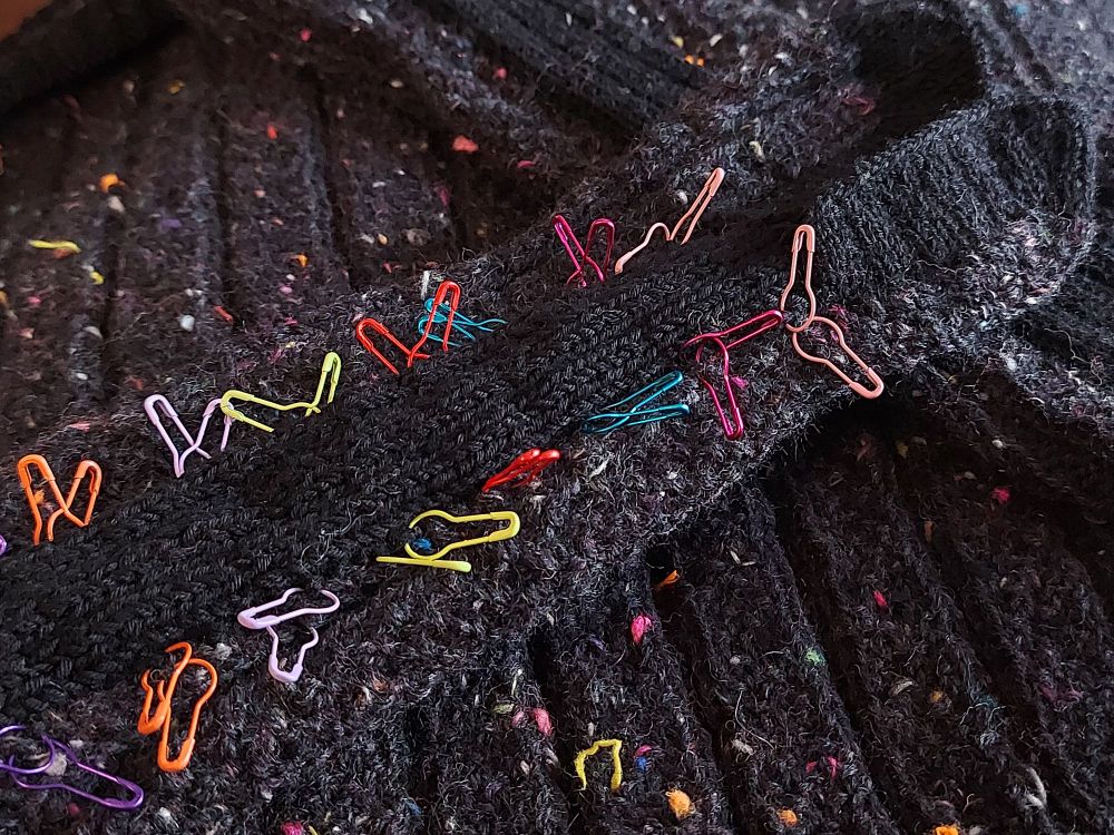 A sleeve is laying on a half finished sweater, with many color coded stitch markers. It almost looks like the back of a dinosaur. Most of the parts are made of  a charcoal colorway covered with lots of colorful blips, hem and intarsia are in solid black.

#knittersofsky #knitting #fiberarts #handmade #memade 