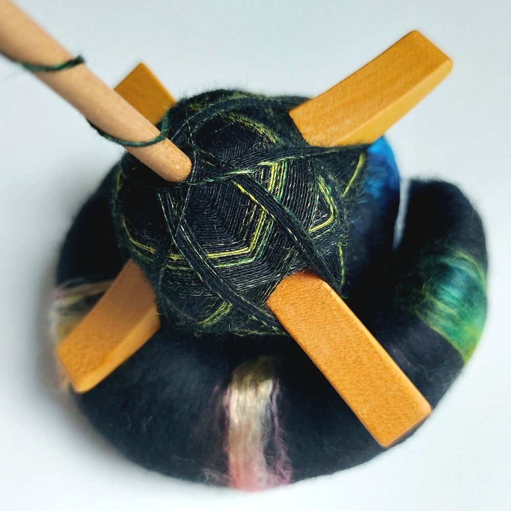 a closeup of a yellowheart crossarm spindle, filled with black yarn and yellow-green-ish streaks, sitting on a black rolag with colorful silk parts