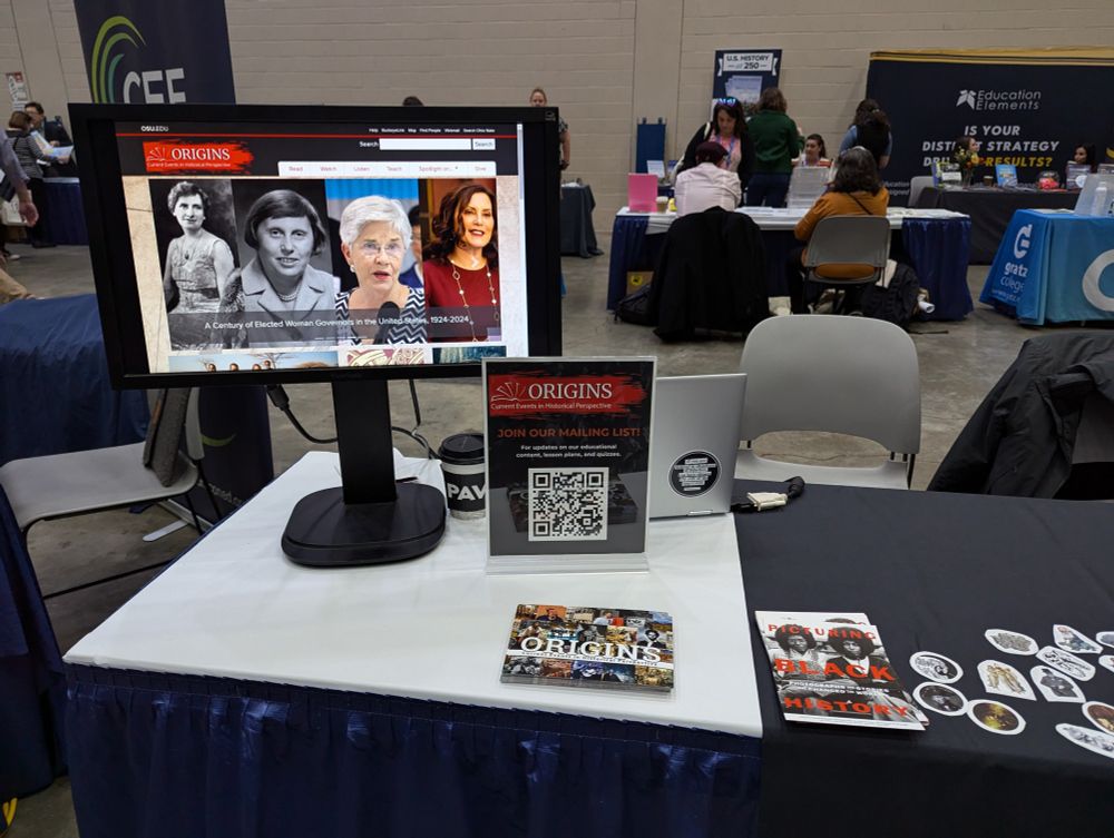 A computer monitor displaying the Origins website on a table with postcards, stickers and a sign with a QR code for joining our email list.