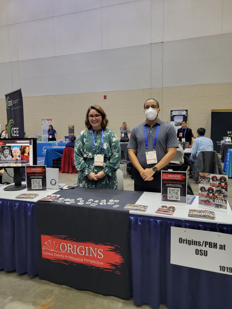 Kristin Osborne and Paul McAllister stand behind table with monitor displaying the Origins website and signage with QR codes to sign up for a mailing list. The table also displays postcards and stickers.