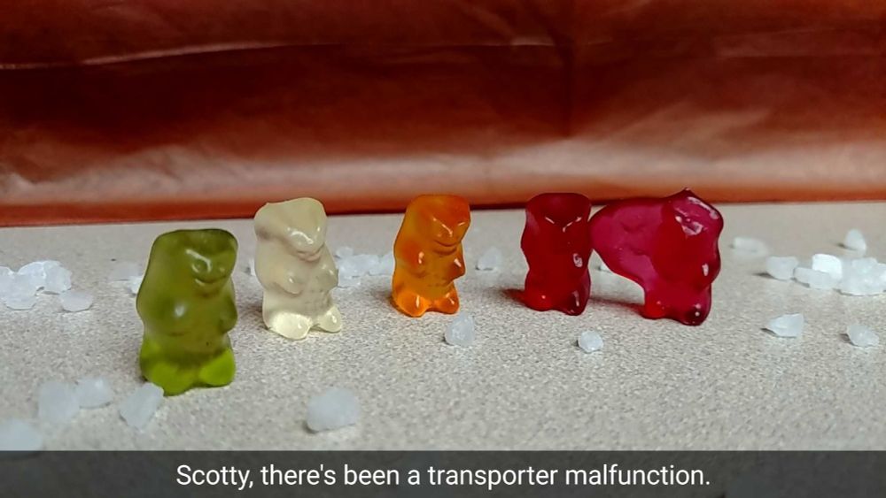 There are five gummy bears (green, white, orange, and two red) standing upright. The red gummy bear all the way on the right is deformed with a large blob coming off it's left side -- the same side touching the other red gummy bear, like they are attached. Large grains of salt (mimicking boulders on another planet) are all around the gummy bears and a brown background (like an alien sky). The caption says, "Scotty, there's been a transporter malfunction." Poor Red Shirt Gummy Bears.