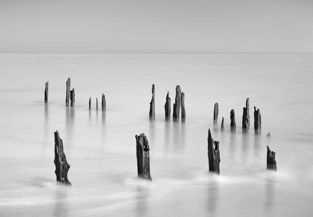Black and white image of two converging lines of wooden posts in shallow outgoing tide. A slow shutter speed smooths the water.